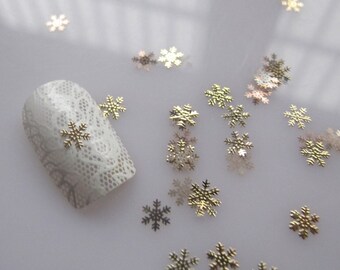 May include: Gold snowflake nail art embellishments scattered on a white surface. A white nail with a lace design has a gold snowflake on it.