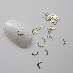 Puede incluir: Adornos para uñas en forma de estrella dorada esparcidos sobre una superficie blanca. Algunas de las estrellas están conectadas en un patrón de cadena.