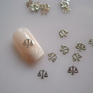Può includere: Ciondoli argentati per nail art a forma di bilancia della giustizia. I ciondoli sono sparsi su una superficie bianca con un ciondolo su un'unghia.