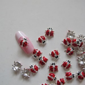 Può includere: Assortimento di ciondoli per unghie a forma di coccinella argentati con smalto rosso e nero. I ciondoli sono sparsi su una superficie bianca con un ciondolo su un'unghia.