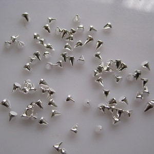 May include: A large quantity of silver-coloured metal pyramid studs. The studs are scattered on a white surface.