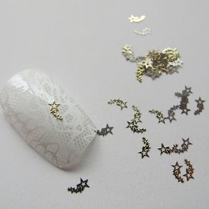 Puede incluir: Una uña blanca con un diseño de estrellas plateadas y doradas. Las estrellas están dispuestas en una línea curva, creando un efecto de estrella fugaz. La uña está rodeada de estrellas plateadas y doradas sueltas.