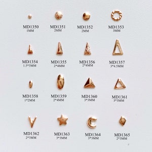 May include: Assortment of gold-toned metal nail art charms in various shapes and sizes, including circles, triangles, stars, moons, and diamonds. The charms are arranged in rows on a white background. Each charm is labelled with a product code and size in millimetres.