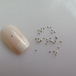 May include: A close-up of a fingernail with a small silver nail stud. Scattered around the nail are many more silver nail studs.
