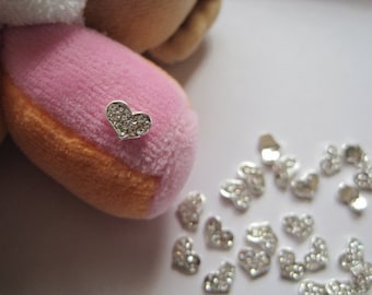 May include: A close-up of a silver heart-shaped charm with rhinestones. The charm is on a pink and orange plush toy. There are many more silver heart charms scattered on a white surface.