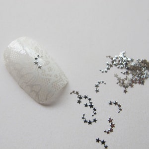 Puede incluir: Una uña blanca con un diseño de encaje y arte de uñas en forma de estrella plateada. También hay piezas sueltas de arte de uñas en forma de estrella plateada esparcidas sobre una superficie blanca.