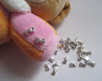 May include: A close-up of a plush toy with a pink and orange body. Two silver bow-shaped charms with rhinestones are attached to the toy. A pile of silver bow-shaped charms with rhinestones are scattered on a white surface.