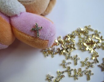 May include: A close-up of a pile of gold-toned cross charms with clear rhinestones. The charms are scattered on a white surface. One charm is also attached to a pink and orange plush toy.