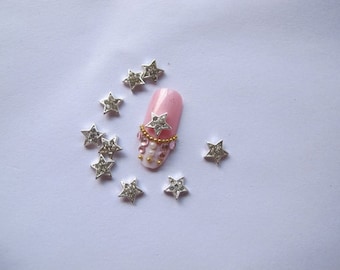 May include: Silver star nail charms with rhinestones. The charms are scattered on a white surface with one charm on a pink nail.