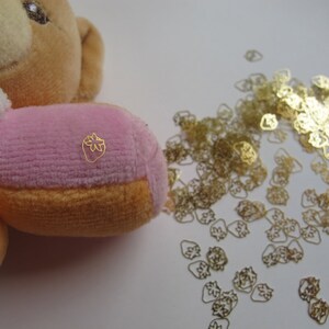 Puede incluir: Un oso de peluche rosa y naranja con un amuleto de fresa dorado en el pecho. El oso está rodeado de una dispersión de amuletos de fresa dorados.