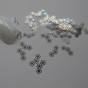 Puede incluir: Una dispersión de pequeñas decoraciones de metal plateado y dorado en forma de flor. Las decoraciones son pequeñas y delicadas, con un delicado diseño calado.