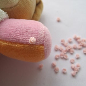 Op de afbeelding: Een roze en oranje pluche speelgoed met een kleine roze roos genaaid op de roze stof. Het speelgoed is omringd door een strooiing van kleine roze rozen.
