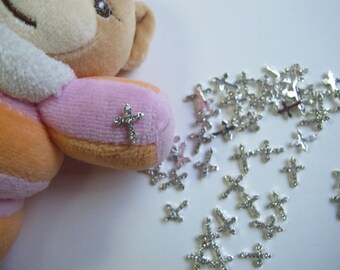 May include: A close-up of a pile of silver rhinestone cross embellishments. The cross embellishments are scattered on a white surface. A pink and orange plush toy is in the background.
