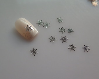 May include: Silver snowflake nail art embellishments on a white background. The snowflakes are scattered around a fingernail with a light pink nail polish.