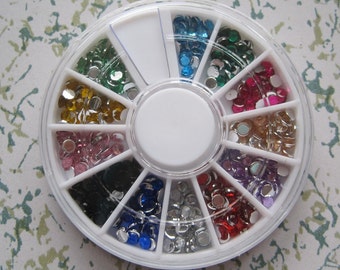 May include: A white plastic wheel with 12 compartments, each filled with a different color of small, round rhinestones. The rhinestones are arranged in a circle around the wheel, with a white center.