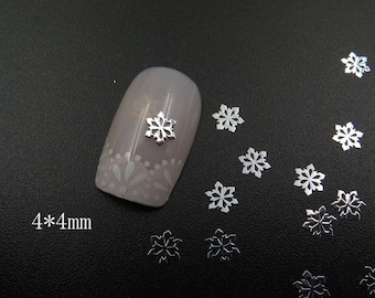 May include: Silver snowflake nail art embellishments, 4mm in size. The image shows a close-up of a fingernail with a silver snowflake on it, and several other snowflakes scattered around.