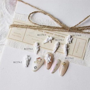May include: White ceramic angel figurines with wings, ideal for nail art. The figurines are arranged on a white background with a piece of twine and a card with the text "ERRY SEED COMPANY CLARINDA RC279-3 (dot was above space) (Wrapper Chaser zone) StreetNo RE RC279-1 1367 Sta Post O. RC279-6 RC279-5 Warehouse RC279-4 RC279-2".