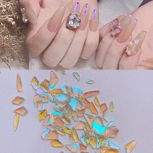Op de afbeelding: Een close-up van iriserende holografische nagelkunst glitter in verschillende vormen en maten. De glitter is verspreid over een wit oppervlak en lijkt van plastic te zijn.