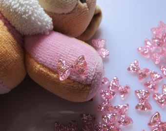 May include: Pink and white plush toy bear with a pink bow on its paw. The bear is surrounded by pink glitter bow embellishments.