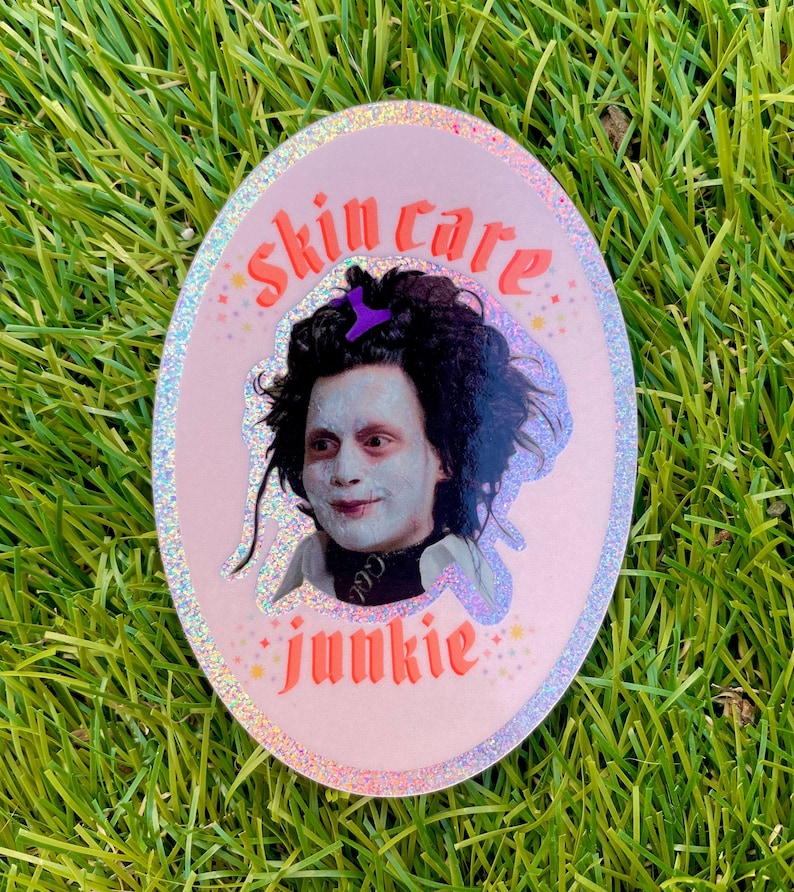 May include: A pink oval sticker with a glittery finish. The sticker features a photo of Johnny Depp as Edward Scissorhands with the text "skin care junkie" in a pink and orange font.
