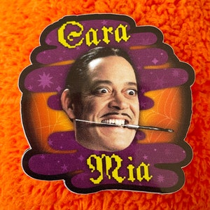 May include: A purple and orange sticker with a cartoon image of a man with a mustache and a paintbrush in his mouth. The text "Cara Mia" is written in yellow letters on the sticker.