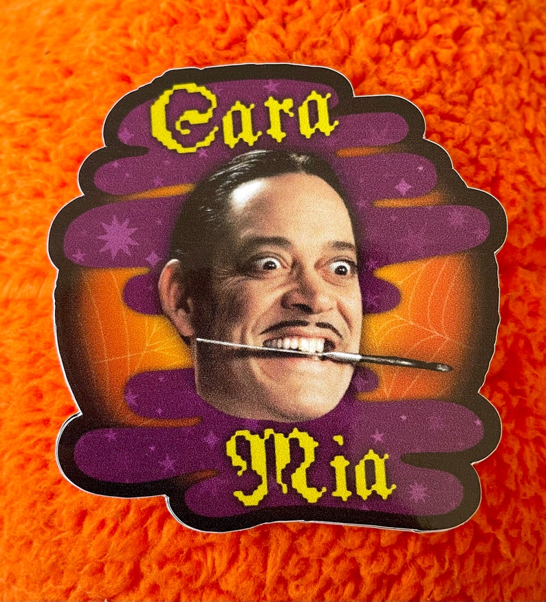 May include: A purple and orange sticker with a cartoon image of a man with a mustache and a paintbrush in his mouth. The text "Cara Mia" is written in yellow letters on the sticker.