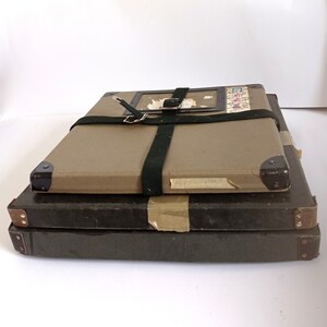 Könnte beinhalten: Drei gestapelte, rechteckige, vintage, braune und schwarze Aufbewahrungsboxen mit einem grünen Canvas-Riemen und einer Schnalle. Die obere Box hat ein kleines, rechteckiges, braunes Papieretikett mit einem verblassten Bild und Text.