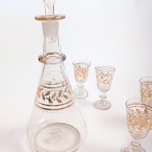 Puede incluir: Un juego de decantador y vasos de chupito de vidrio transparente con detalles en pan de oro. El decantador tiene un tapón y una base ancha. Los vasos de chupito tienen un tallo y un cuenco redondo.