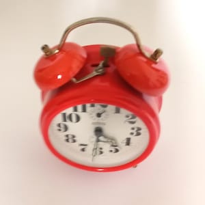May include: A bright red vintage alarm clock with a white clock face. The clock has a metal handle and two red bells on top. The clock face displays black numbers and hands.
