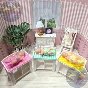 May include: Miniature nursery scene with three baby dolls on changing tables. Each doll wears a crocheted outfit in pink, green, and yellow. The room has pink and white striped walls, a window, and a small white dresser.
