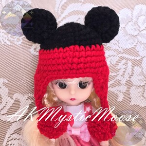 May include: A red and black crocheted hat with Mickey Mouse ears. The hat has a pom-pom on each ear and a pom-pom on the end of each flap.
