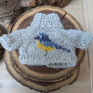 May include: A small grey knitted jumper with a blue and yellow bird design. The jumper has a turtleneck and is made of a soft, fluffy yarn.