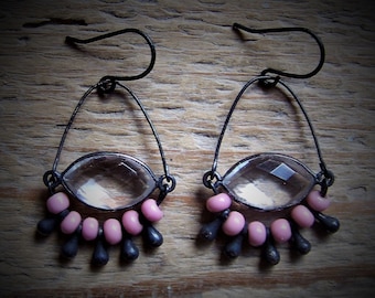 May include: A pair of handcrafted earrings with a clear, faceted oval stone set in a dark metal frame. Pink beads and dark teardrop-shaped accents dangle below, suspended from dark metal ear wires. The earrings are displayed on a wooden surface.