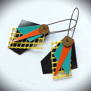Puede incluir: Pendientes colgantes geométricos con una combinación de colores negro, turquesa, naranja y amarillo. Los pendientes tienen un gancho de alambre negro y un acento circular de color latón. El diseño incluye un trapecio negro, un cuadrado turquesa y un triángulo naranja.