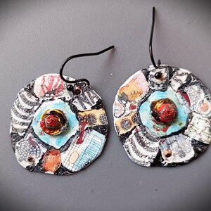 May include: A pair of handmade, circular earrings. Each earring features a central red and yellow design surrounded by a light blue circle. The outer edges are a mix of black, white, and colorful patterned pieces, with black wire hooks.