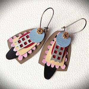 Puede incluir: Un par de pendientes abstractos con un diseño único. Los pendientes presentan una combinación de formas y colores, incluyendo un círculo azul claro, un cuadrado rojo con una rejilla plateada y una base marrón con acentos rosas y negros. Los pendientes tienen ganchos plateados y detalles dorados.