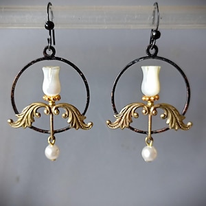 Può includere: Un paio di orecchini pendenti con una cornice circolare in metallo scuro, un bocciolo centrale bianco madreperlaceo, dettagli a foglia dorata decorati e una piccola perla sferica bianca appesa a ganci neri per le orecchie.