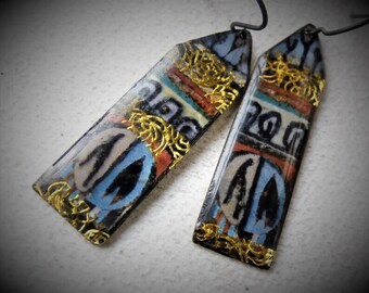 May include: A pair of long, rectangular earrings with a black, white, blue, and gold geometric pattern. The earrings are made of resin and have a gold-tone finish.