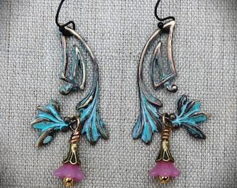 May include: A pair of artisan earrings with a botanical theme. The earrings feature a bronze-colored, leaf-shaped design with turquoise accents. Each earring has a small, pink, bell-shaped charm with a gold-colored bead.