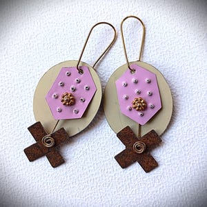 Puede incluir: Pendientes hechos a mano con un hexágono rosa claro, tachuelas plateadas y una flor dorada, unidos a una cruz marrón y un gancho de alambre dorado. Los pendientes están colocados sobre un fondo ovalado crema.