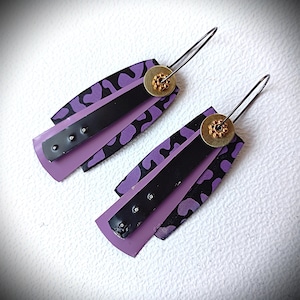 Puede incluir: Un par de pendientes abstractos con un diseño en capas. Cada pendiente presenta una base de color lavanda, una tira central negra y una capa superior con estampado morado y negro. Un pequeño disco dorado con un diseño floral en la parte superior.