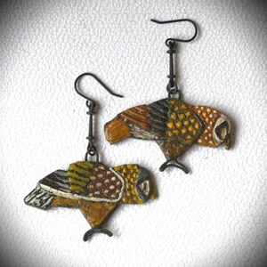 Könnte beinhalten: Ein Paar Hängeohrringe mit handbemalten Eulenmotiven. Die Eulen sind in Braun-, Gelb-, Weiß- und Schwarztönen bemalt. Die Ohrringe sind mit Metallhaken gefertigt.