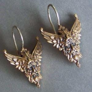 May include: A pair of gold-colored earrings featuring a winged face design. The earrings have a detailed, antique appearance with intricate facial features and wing details. The earrings are suspended from silver-colored ear wires.