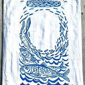 May include: White kitchen towel with a blue linocut print of fish swimming in the ocean. The design includes stylized waves, fish, and a circular shape. The towel is made of a soft, absorbent material.