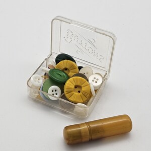 May include: A clear plastic box filled with various colorful buttons, including yellow, green, white, and black. A small, cylindrical, amber-colored container rests beside the box. The buttons are of different sizes and shapes.