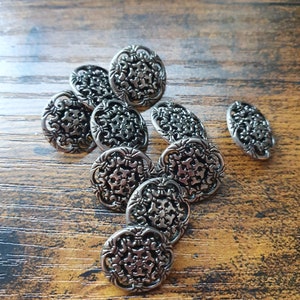 Könnte beinhalten: Eine Gruppe silberfarbener Metallknöpfe mit filigranen Blumenmustern. Die Knöpfe liegen lose auf einer Holzoberfläche.