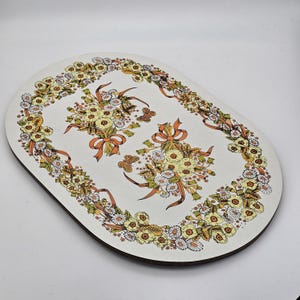 Può includere: Vassoio ovale con un design floreale. Il vassoio presenta uno sfondo bianco con un bordo di fiori gialli e bianchi. Due bouquet con nastri arancioni e farfalle sono al centro. Il vassoio ha un bordo marrone scuro.