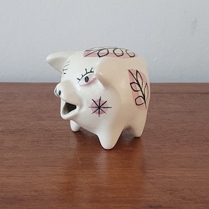 May include: White ceramic piggy bank with pink and black geometric designs. The piggy bank has a stylized face with black eyes and a pink nose.