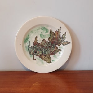Op de afbeelding: Een wit keramiek bord met een handgeschilderd ontwerp van een groene en bruine vis met een groot oog. De vis zwemt in een groene en witte aquarel achtergrond.