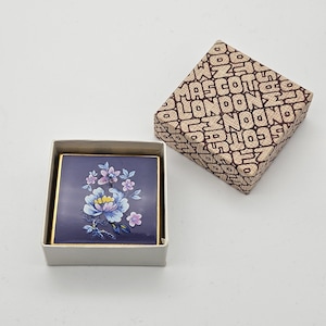 May include: A vintage square compact with a floral design in a white box. The compact has a navy blue background with a gold trim. The box has a patterned lid with the word "LONDON" repeated.